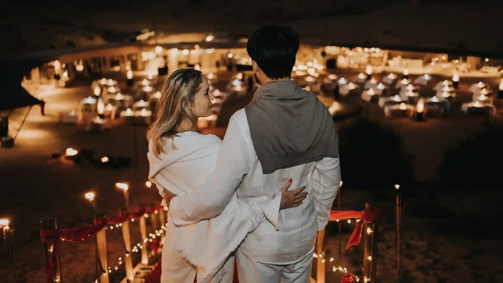Couple embracing while overlooking a candlelit romantic desert dining setup at night.